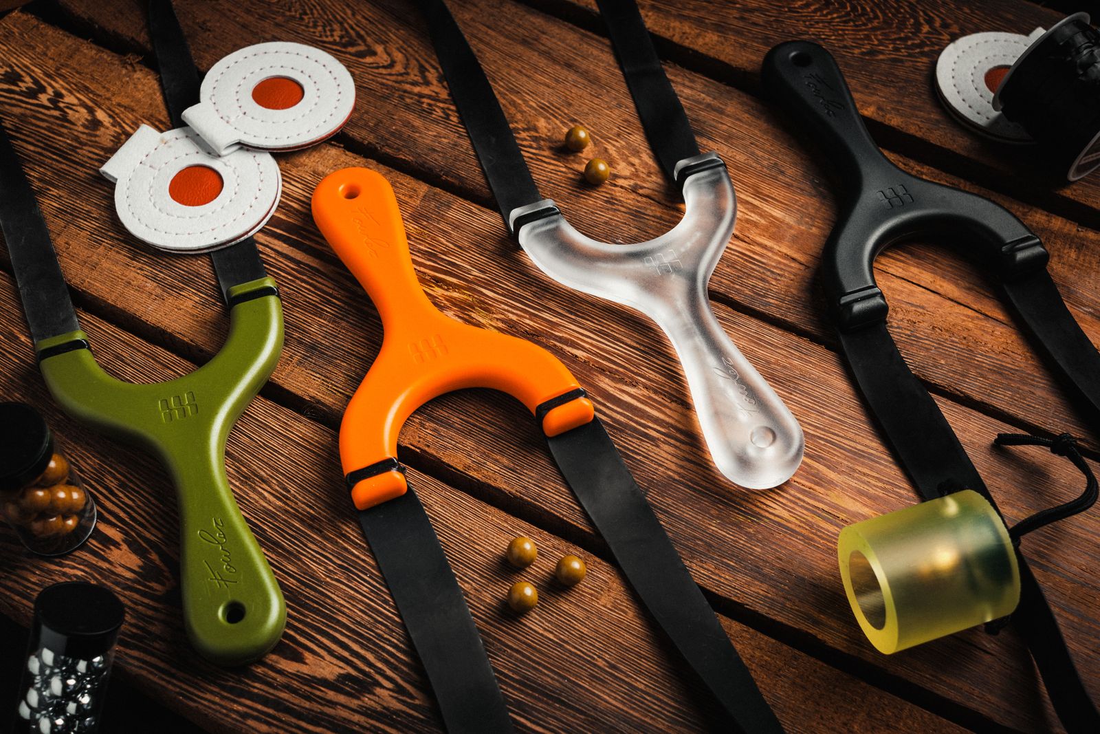 Four Hornet slingshots in green, orange, clear and black laid out on a wooden table with bands, targets and ammo