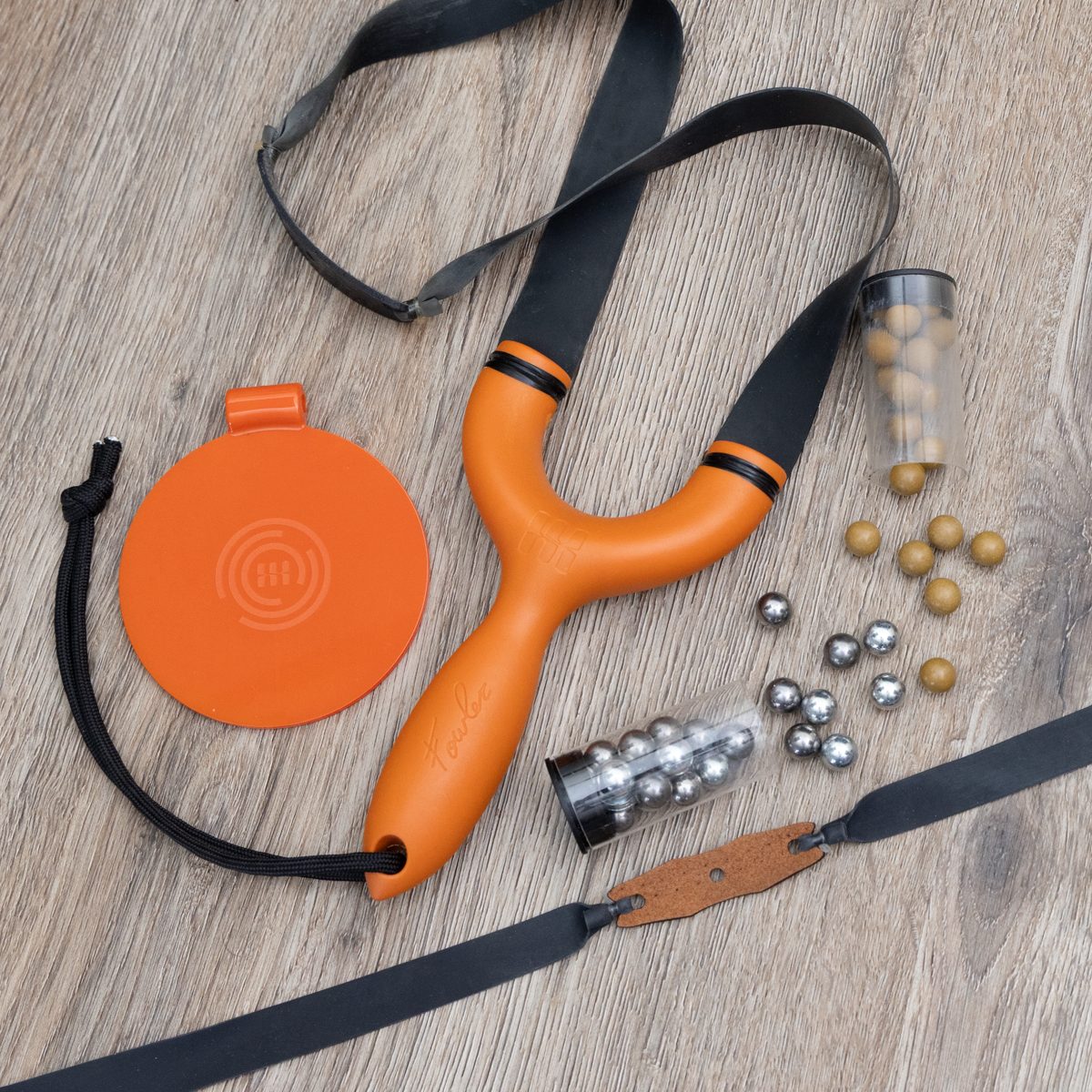 An orange Sparrow slingshot laid out on wood with two bandsets, an orange spinner target, clay and steel ammo and a paracord lanyard