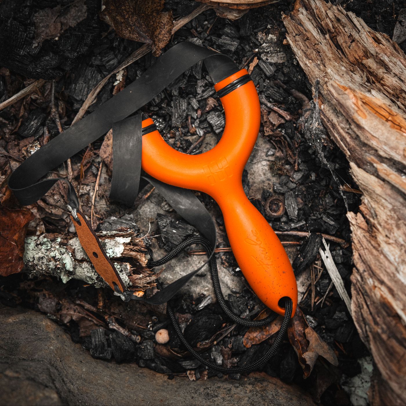 An orange Sparrow slingshot with its bands and paracord lanyard on a dark forest floor beside a weathered log
