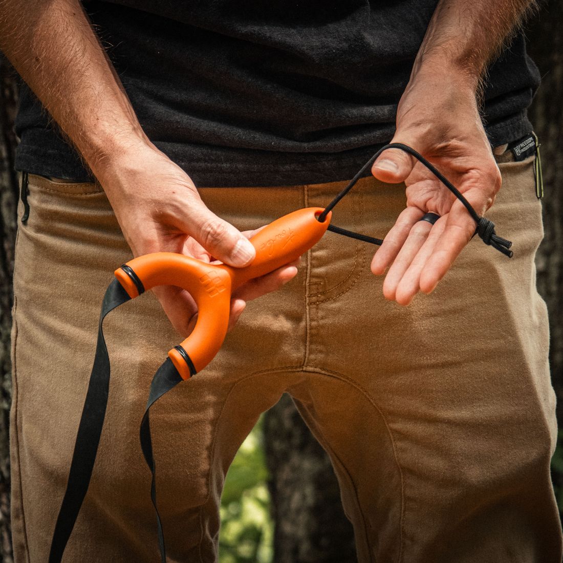 A man in khakis holding an orange Sparrow slingshot, its paracord lanyard looped over his other hand