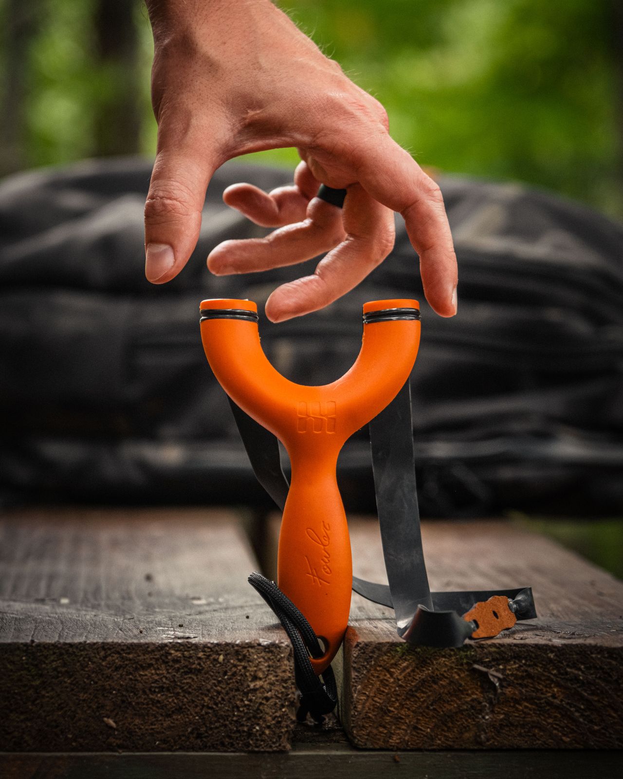 A hand reaching for an orange Sparrow slingshot standing upright between two weathered boards, bands hanging, woods behind