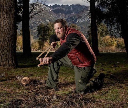 Zach Fowler kneeling in a clearing with a slingshot in his hands, mountains behind him